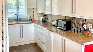 Eine moderne Küche mit weißen Schränken, einem Geschirrspüler, einer Mikrowelle und einem Toaster. Ein Fenster mit Jalousien und eine Uhr sind an der Wand angebracht.
