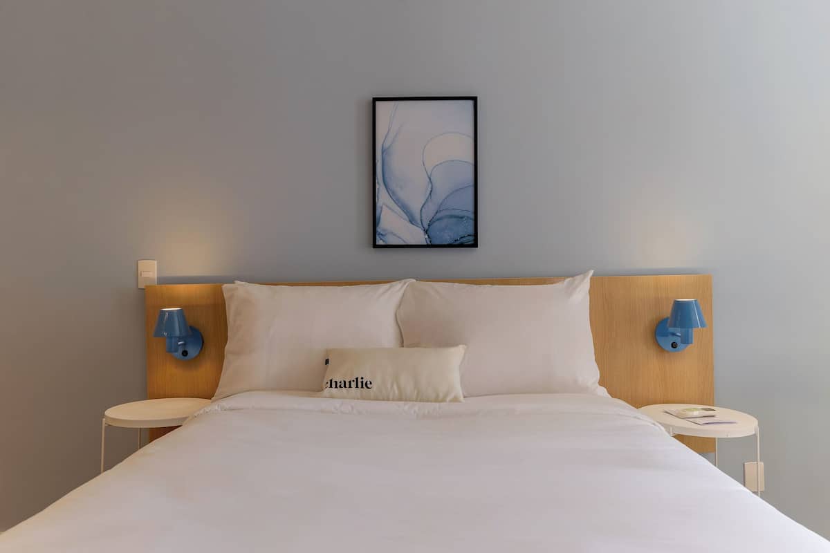 Una cama bien hecha con una almohada que dice 'charlie', dos lámparas fijadas a la pared y un cuadro abstracto enmarcado sobre la cama.