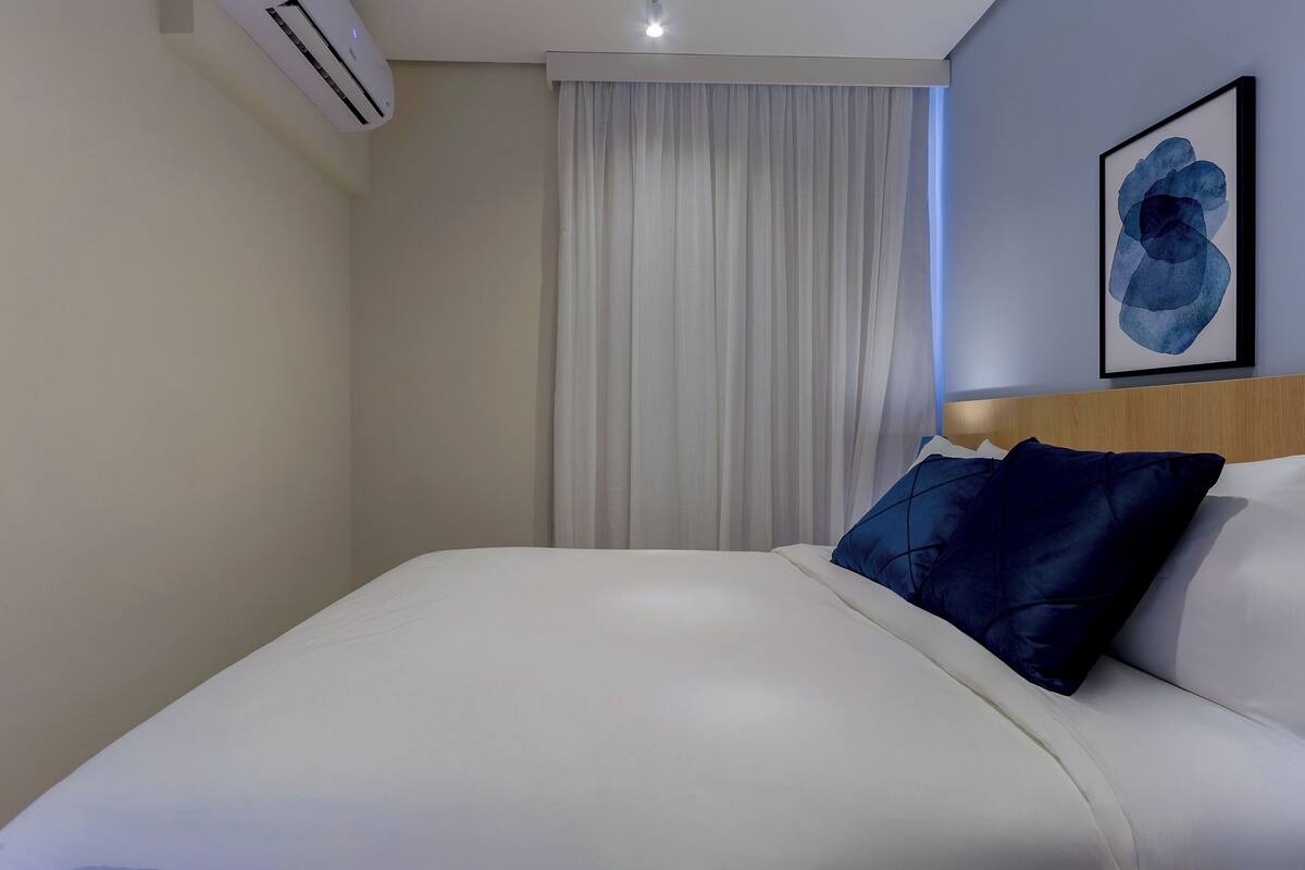 Habitación de hotel con cama, cabecera, aire acondicionado instalado en la pared, ventana con cortinas y una obra de arte enmarcada.