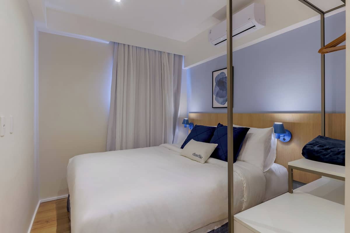 Una habitación de hotel con cama, mesita de noche, aire acondicionado instalado en la pared y cortina.