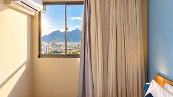 Une chambre d’hôtel avec un lit, une fenêtre avec des rideaux, un appareil de climatisation et une vue sur un paysage urbain avec des montagnes.
