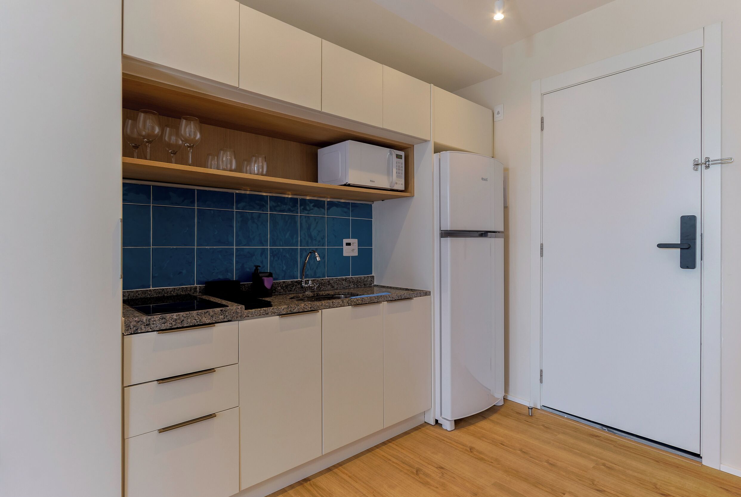 Una cocina moderna con armarios blancos, salpicadero azul, microondas, refrigerador y estante de madera con vasos.