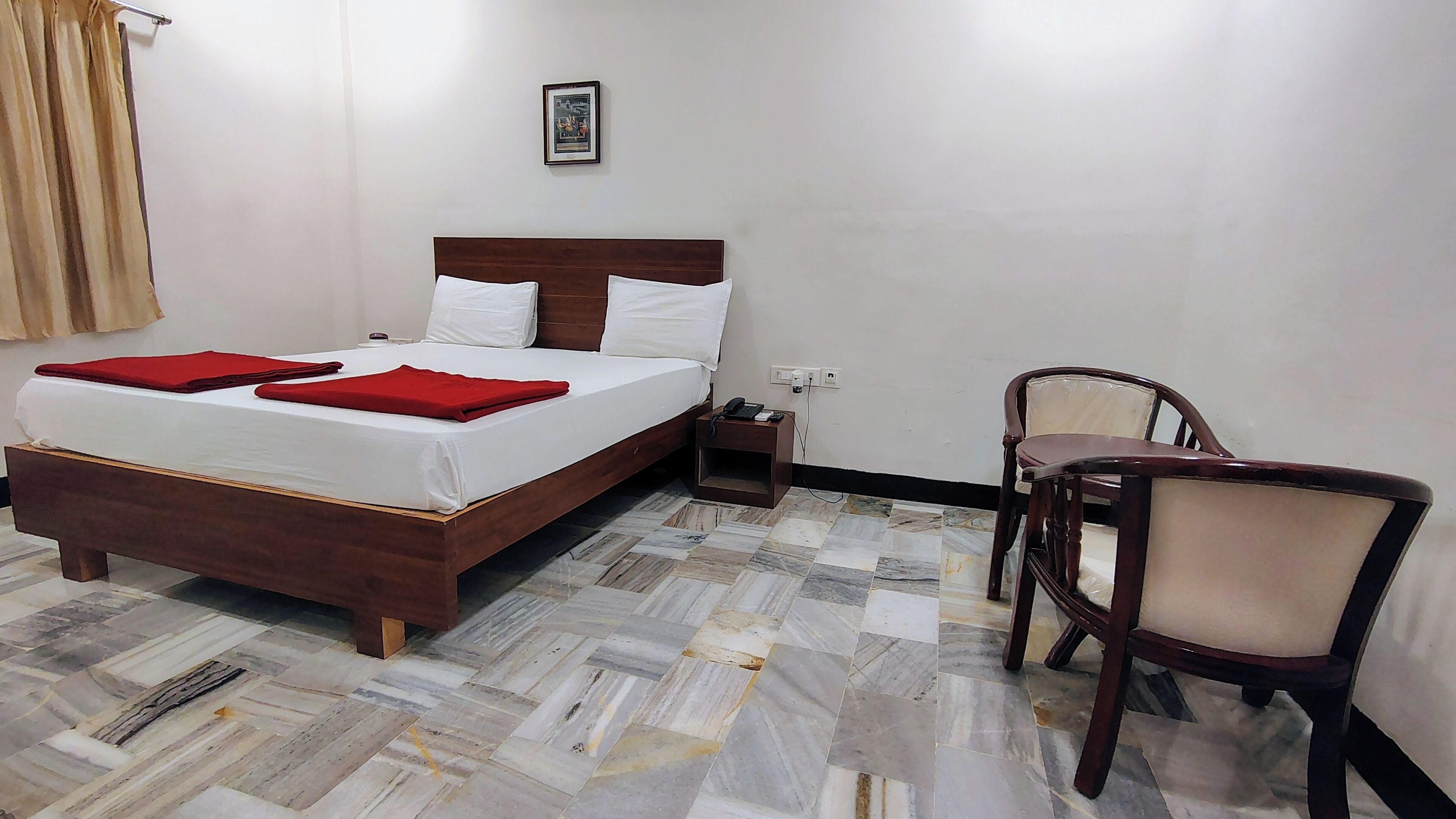 Habitación de hotel con cama de madera, ropa de cama blanca, una manta roja, una mesita de noche pequeña con un teléfono y una silla.