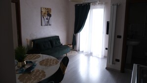 Una sala de estar con un sofá, una mesa de comedor con sillas, un cuadro en la pared y un ventanal con cortinas.