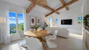 A modern living room with a round wooden table, white chairs, a white sofa, a TV mounted on the wall, and a view of the landscape through large windows. - Charming villa - stylish, quiet, relaxing ***New at FeWo*** (Cavalaire-sur-Mer)
