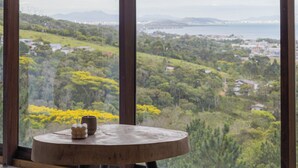A room with a round wooden table, a green cushion, and a view of a landscape through large windows.
