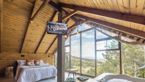A bedroom with a large bed, a view of the outdoors, a wooden ceiling, and a mounted television.