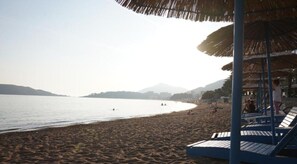 A beach with lounge chairs and umbrellas, a calm sea, and distant mountains. - Modern, Bright Beachfront Flat in Rafailovici (Rafailovići, Opština Budvag, Apartment 2, Bečićka Plaza, 85310, Montenegro)
