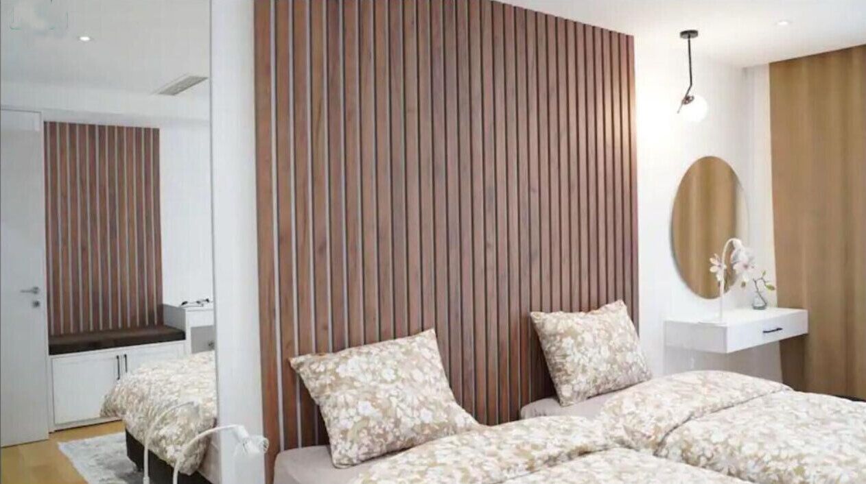 A modern bedroom with a large bed, a mirror, a bedside table, and a wooden panel wall.