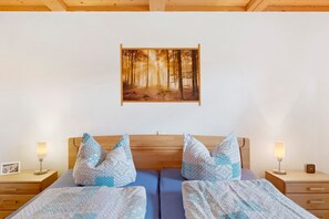 Een slaapkamer met een houten hoofdeinde, twee bedden met beddengoed met een patroon, nachtkastjes met lampen, en een lijst met daarop een boslandschap.