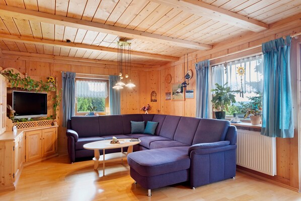 Een houten hutje aan de binnenkant, met een hoekbank, een televisie en een eethoek.