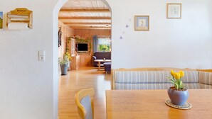 A dining area with a wooden table, a blue vase with yellow flowers, a striped sofa, and a wooden archway leading to a kitchen. - Apartment Sommerruhe in Jostal near Titisee (Titisee-Neustadt)