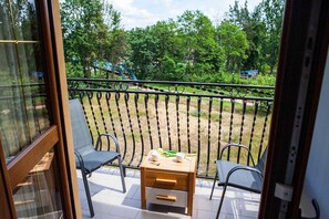 A balcony with two chairs, a small table, and a view of a park with trees and a playground.