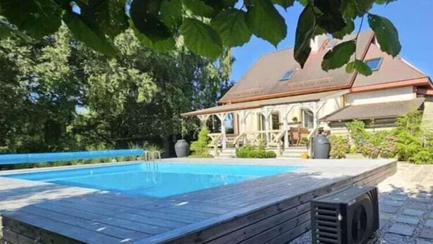 Una piscina con bordo in legno, una veranda coperta e un edificio con tetto spiovente sullo sfondo.
