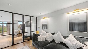En moderne stue med en mørkegrå sofa, hvide puder med mønster, en glasvæg og et stort abstrakt billede på væggen.