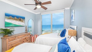 Ein Schlafzimmer mit zwei Betten, einem Deckenventilator, einem großen Fenster mit Meerblick, einer hölzernen Kommode und einem Bild an der Wand.
