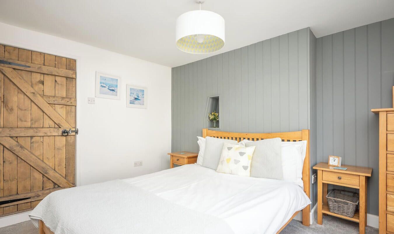 Een slaapkamer met een houten bed, nachtkastje, wandspiegel en een houten kledingkast.