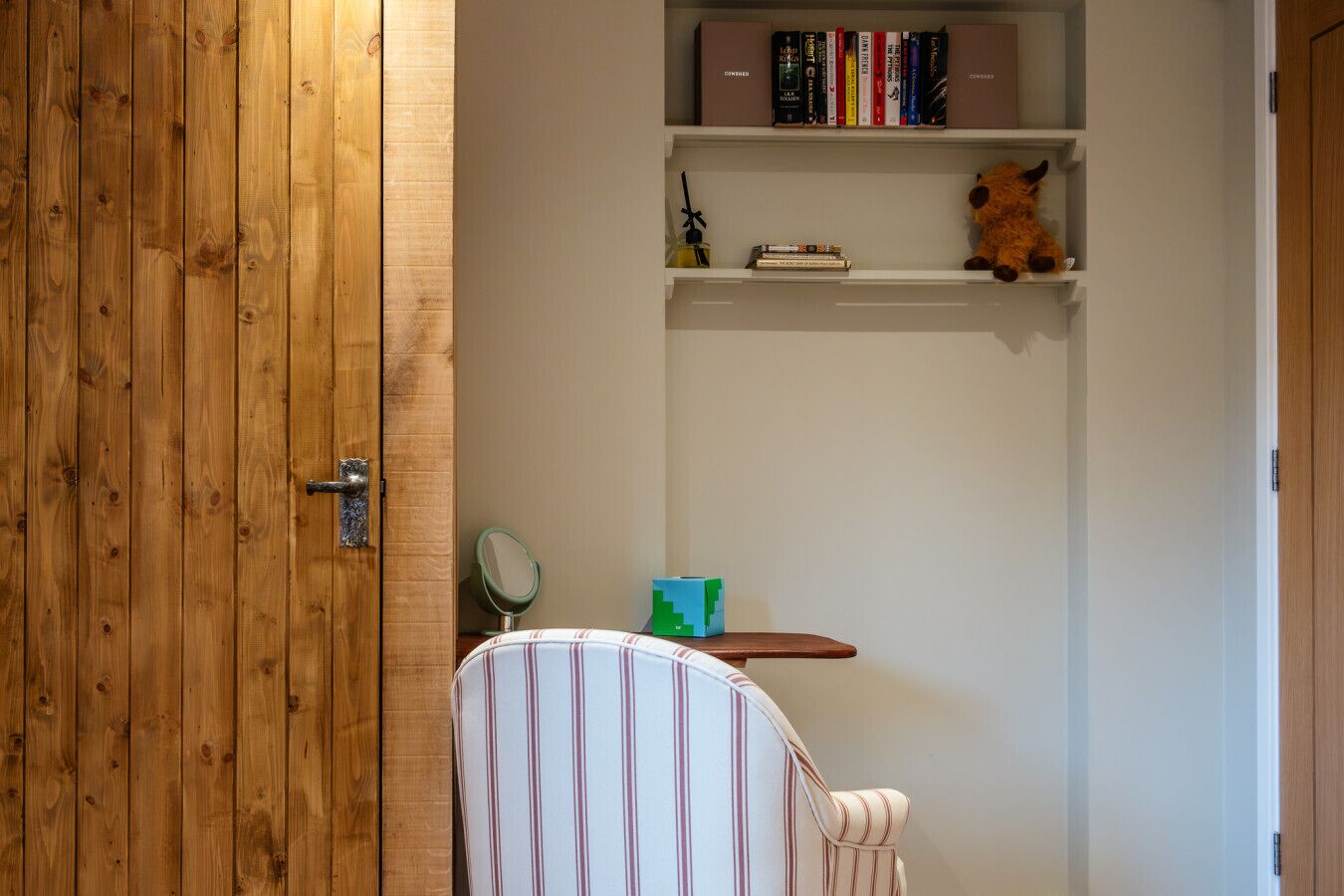 A wooden door, a striped chair, a small table with a mirror, and a shelf with books and a stuffed toy.