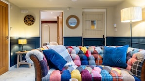 Een gezellige woonkamer met een kleurrijke, gestoffeerde bank, een klein bijzettafeltje met een lamp, en een deur naar een andere kamer.