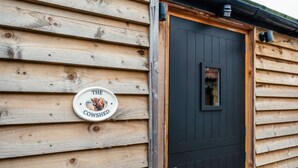 Een houten blokhut met een zwarte deur, een klein raam en een bordje waarop staat 'The Cow Shed'.
