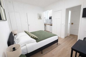 Ein Schlafzimmer mit Bett, Nachttisch, Lampe und Blick zur Küche.