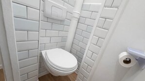 A small, white-tiled bathroom with a toilet, a white pipe, and a roll of toilet paper on the wall. - Charmant Studio Paris 15eme - Proche Tour Eiffel (Paris)