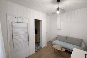 Ein Zimmer mit grauem Sofa, einem Holztisch, einem Wäscheständer aus Metall und einer Deckenleuchte.