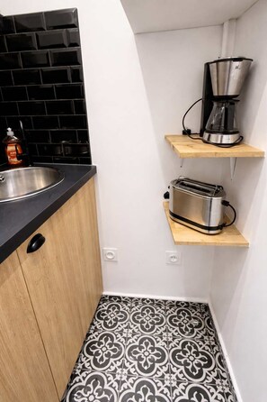Eine kompakte Küche mit schwarzem Fliesenrückwand, einem Edelstahl-Spülbecken, einer Kaffeemaschine und einem Toaster auf Holzregalen.