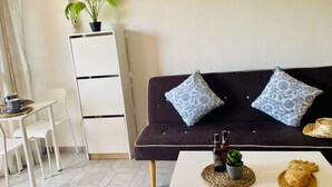 A room with a dark sofa, two patterned pillows, a small table with a plant and two bottles, a white chair, and a potted plant on a cabinet. - Estudio Minerva Splash by At Home Costa del Sol (Benalmádena)