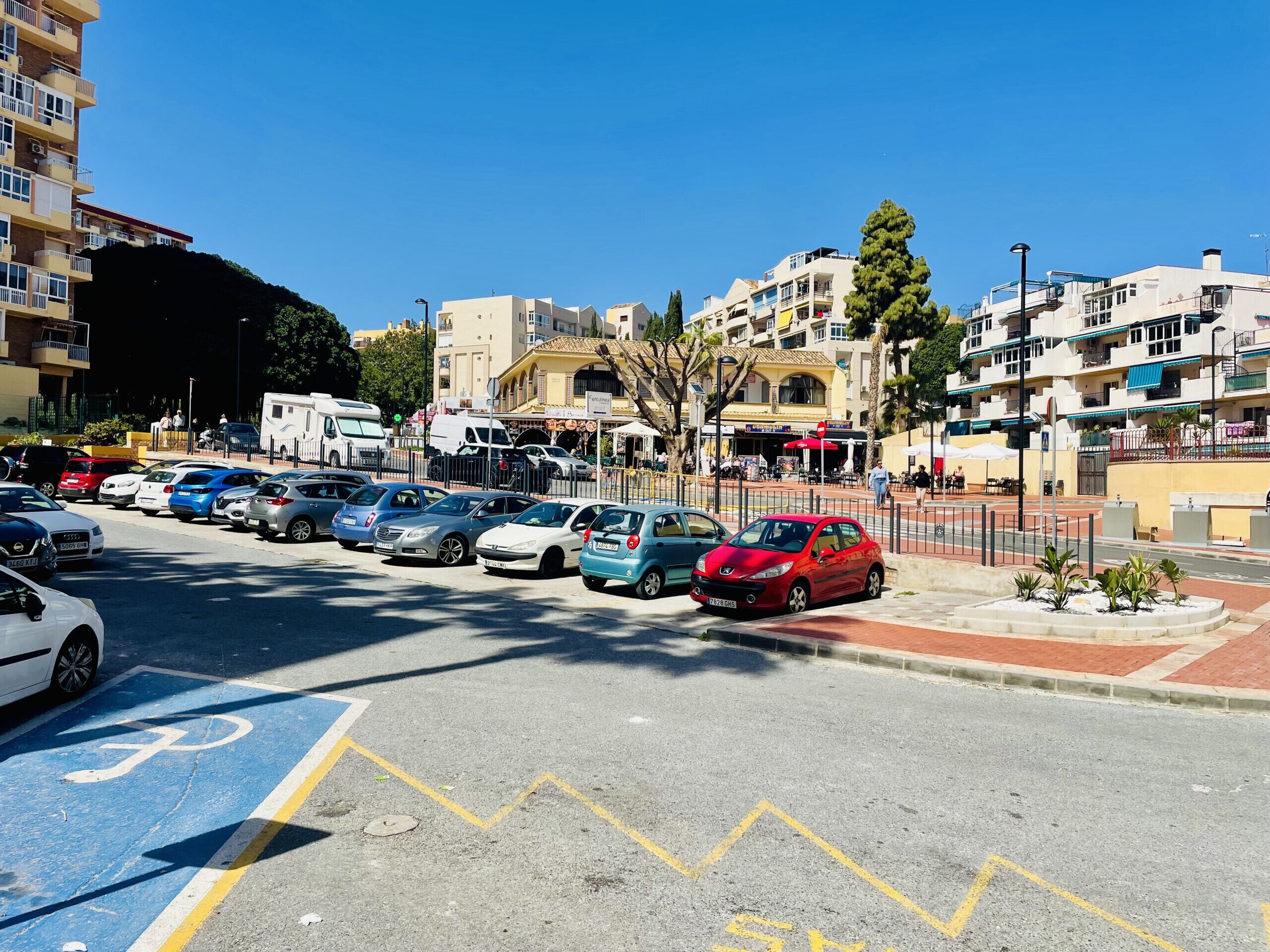 Un parking rempli de voitures, une piste cyclable et, au fond, des immeubles avec des balcons et des boutiques.