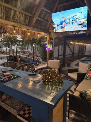 An outdoor seating area with a thatched roof, string lights, and a TV screen displaying a sports event.