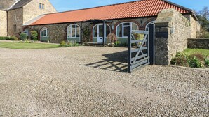 A stone building with a red-tiled roof, a gravel driveway, and a gated entrance. - Castle View Cottage (Bedale)