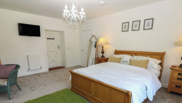A bedroom with a bed, a green rug, a chandelier, a TV, and framed pictures on the wall. - Castle View Cottage (Bedale)