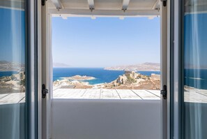 Une vue depuis une pièce dotée d’une grande fenêtre donnant sur un paysage côtier, avec des bâtiments et la mer.