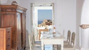 A dining area with a table set for four, a wooden cabinet, and a view of a coastal landscape. - Aegean stones, Ornos, Mykonos (Agios Ioannis Mykonos)