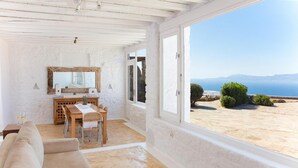 A room with a white interior, a wooden table, a sofa, and a view of the sea. - Aegean stones, Ornos, Mykonos (Agios Ioannis Mykonos)