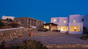 A stone building with a stone staircase, a white building with large windows, and a clear sky. - Aegean stones, Ornos, Mykonos (Agios Ioannis Mykonos)