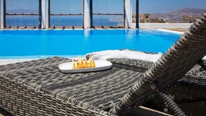 A poolside area with a wicker lounge chair, a plate with a dessert, and a view of the sea and buildings. - Aegean stones, Ornos, Mykonos (Agios Ioannis Mykonos)