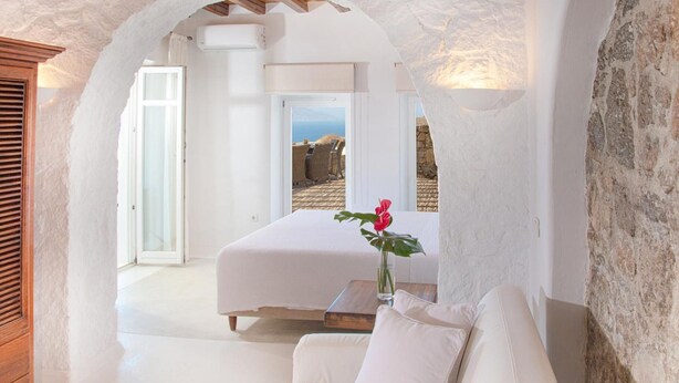A room with a bed, a sofa, a wooden table, and a vase with a flower. - Aegean stones, Ornos, Mykonos (Agios Ioannis Mykonos)