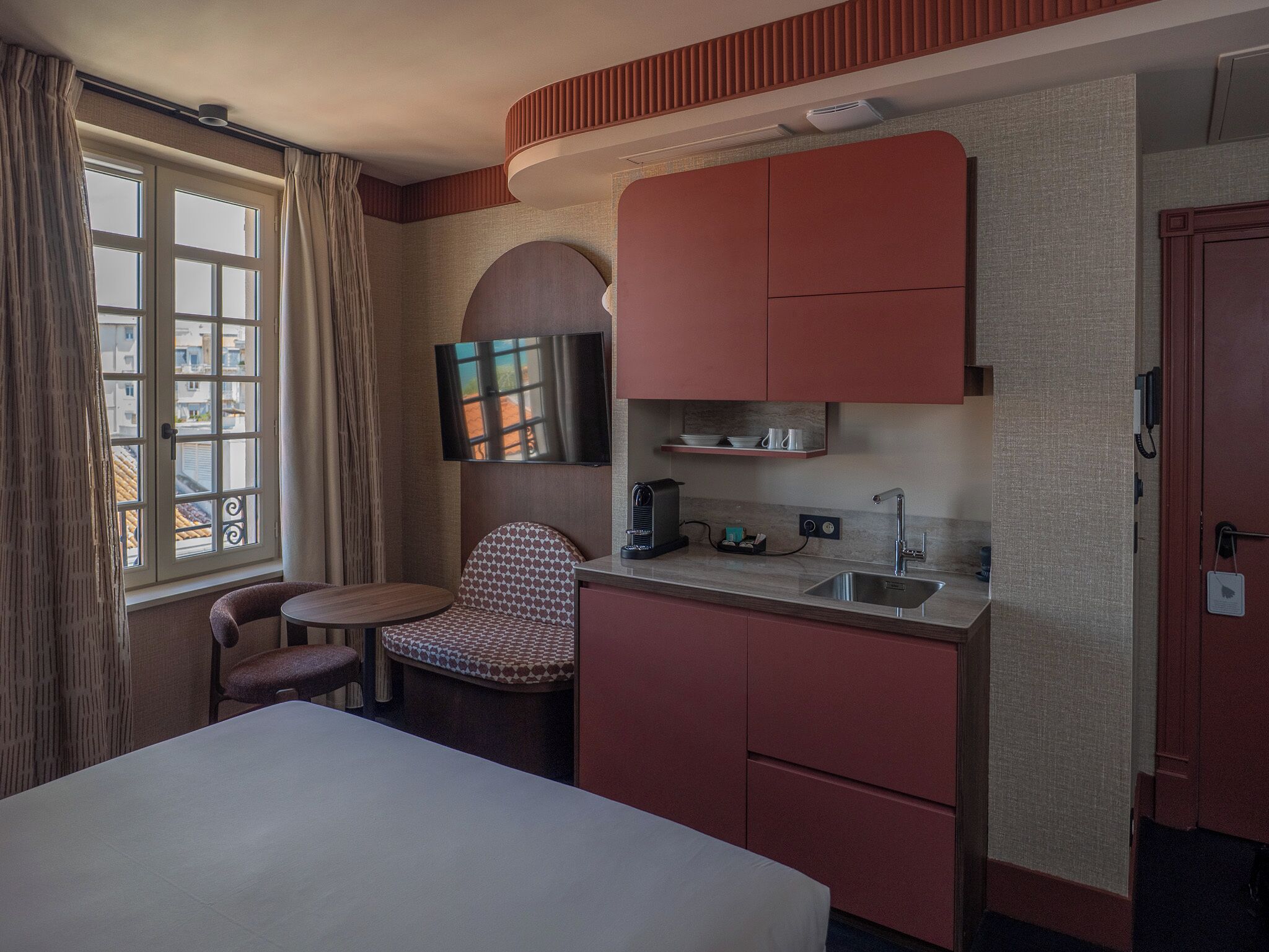 Habitación de hotel compacta con cama, kitchenette pequeña, zona de estar con una silla y televisor montado en la pared.