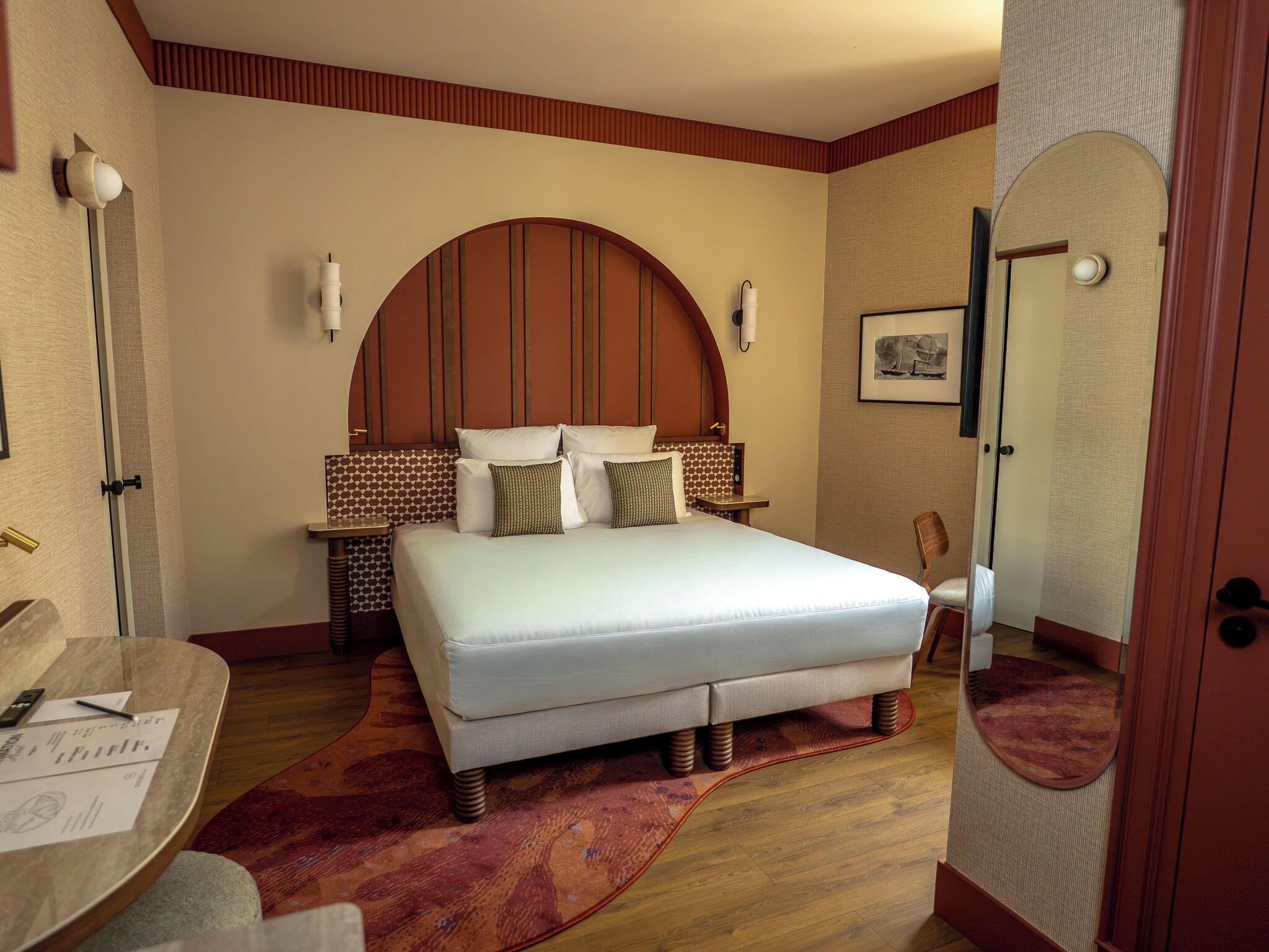 A hotel room with a large bed, a desk with a chair, a mirror, and a door.