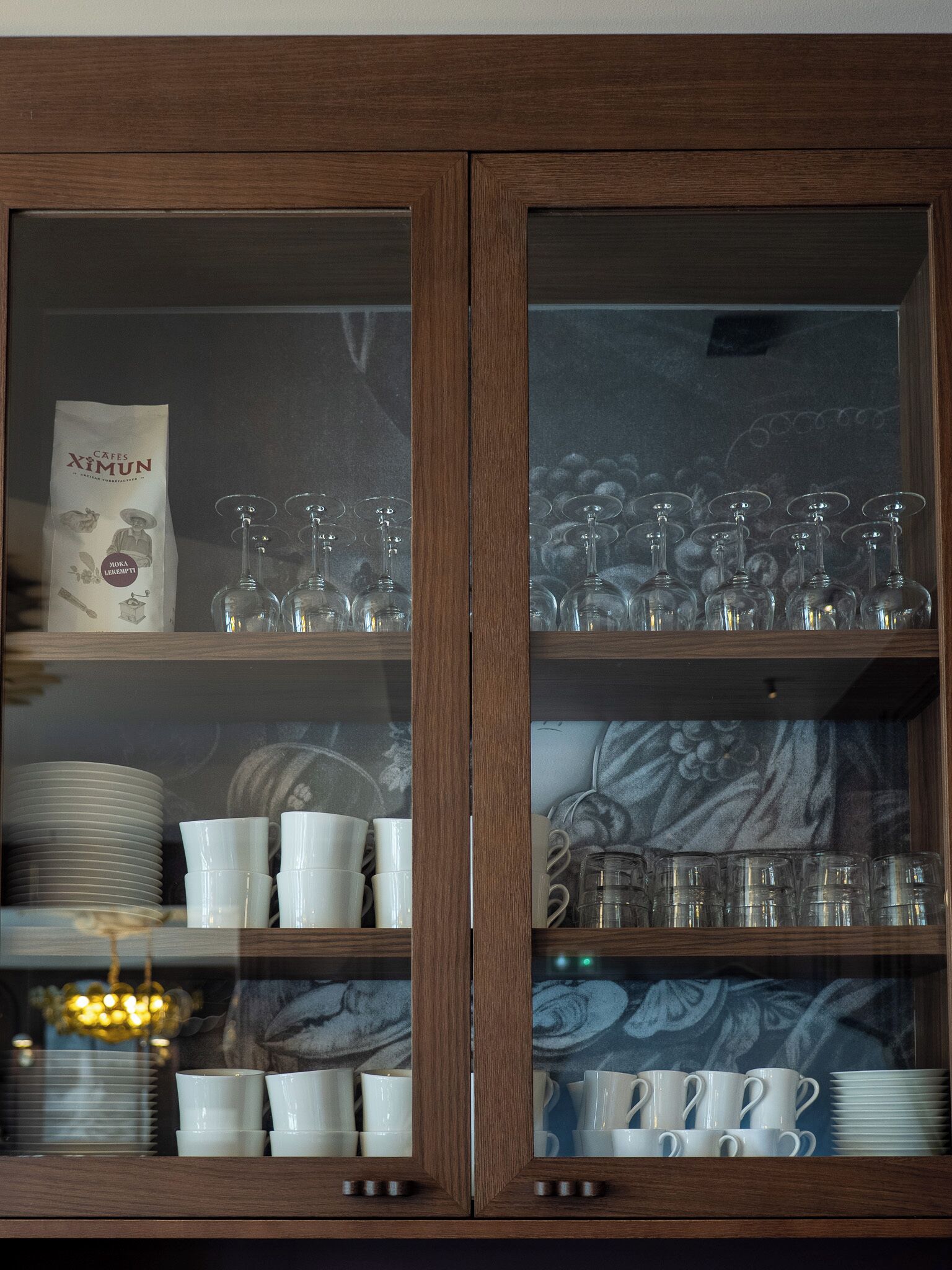 Un armario de madera con puertas de vidrio que contiene tazas, vasos y platos blancos.