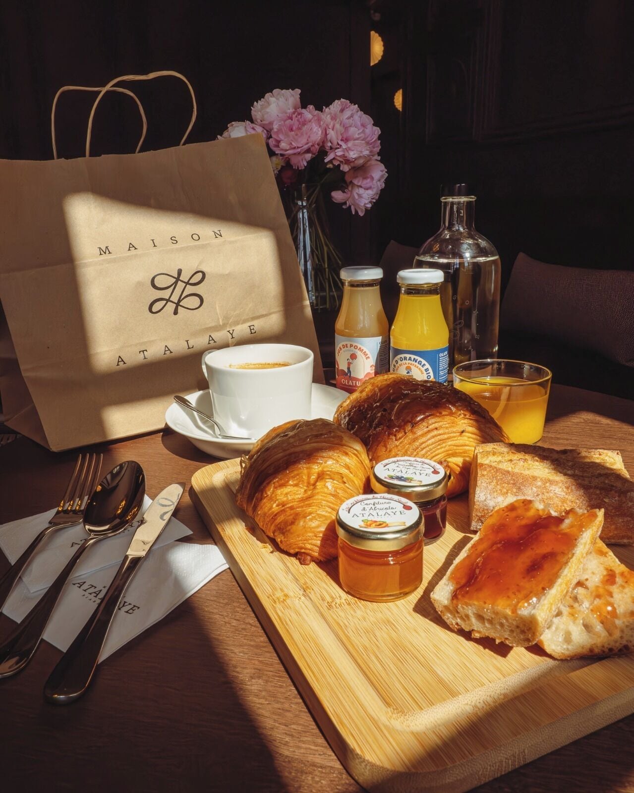 Una mesa de madera con cruasanes, frascos de miel y botellas de jugo, una taza blanca y una bolsa de papel con el texto 'MAISON ATALAYE'.