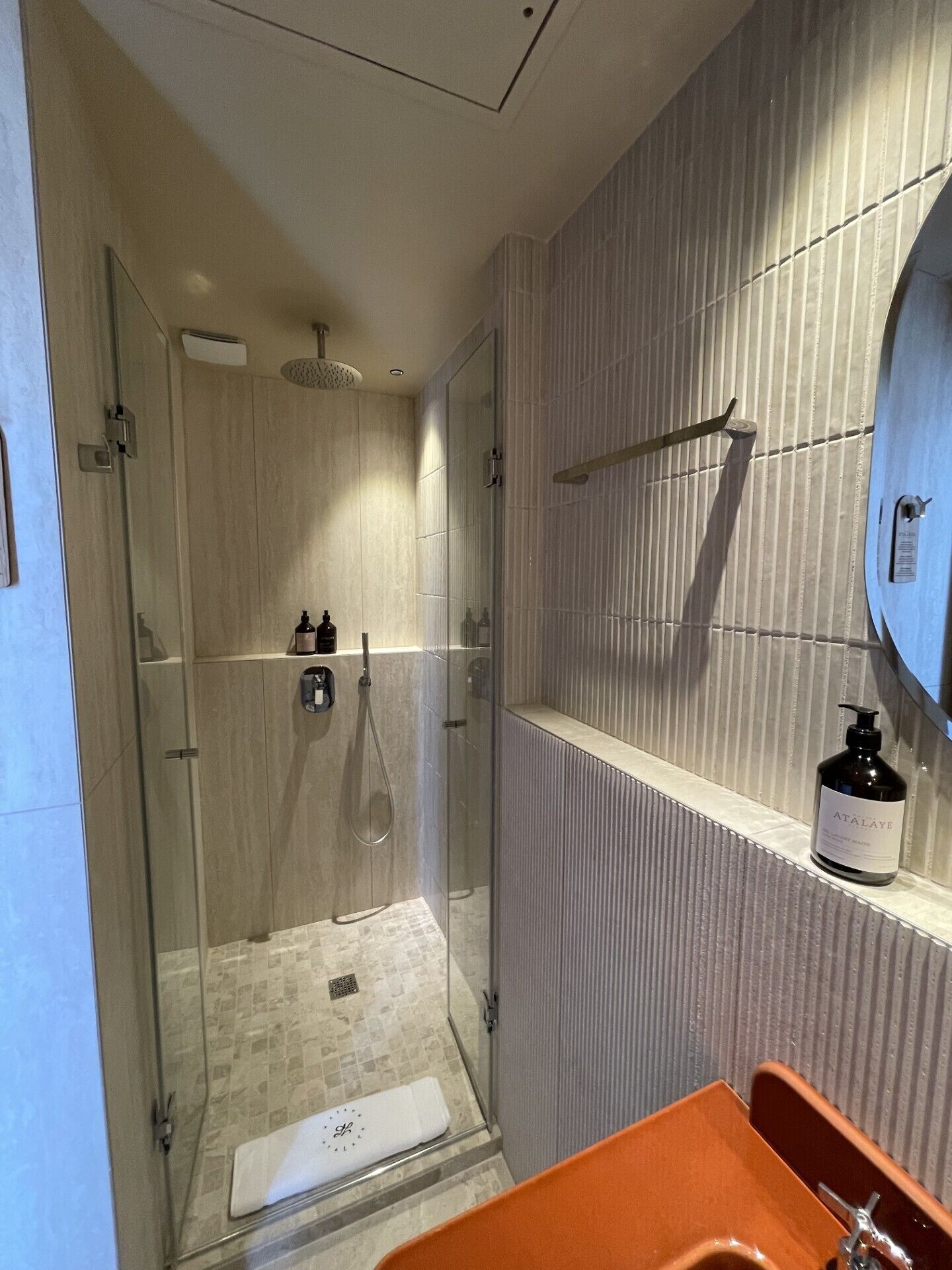 Un baño moderno con ducha de vidrio, repisa empotrada con artículos de aseo y espejo redondo.