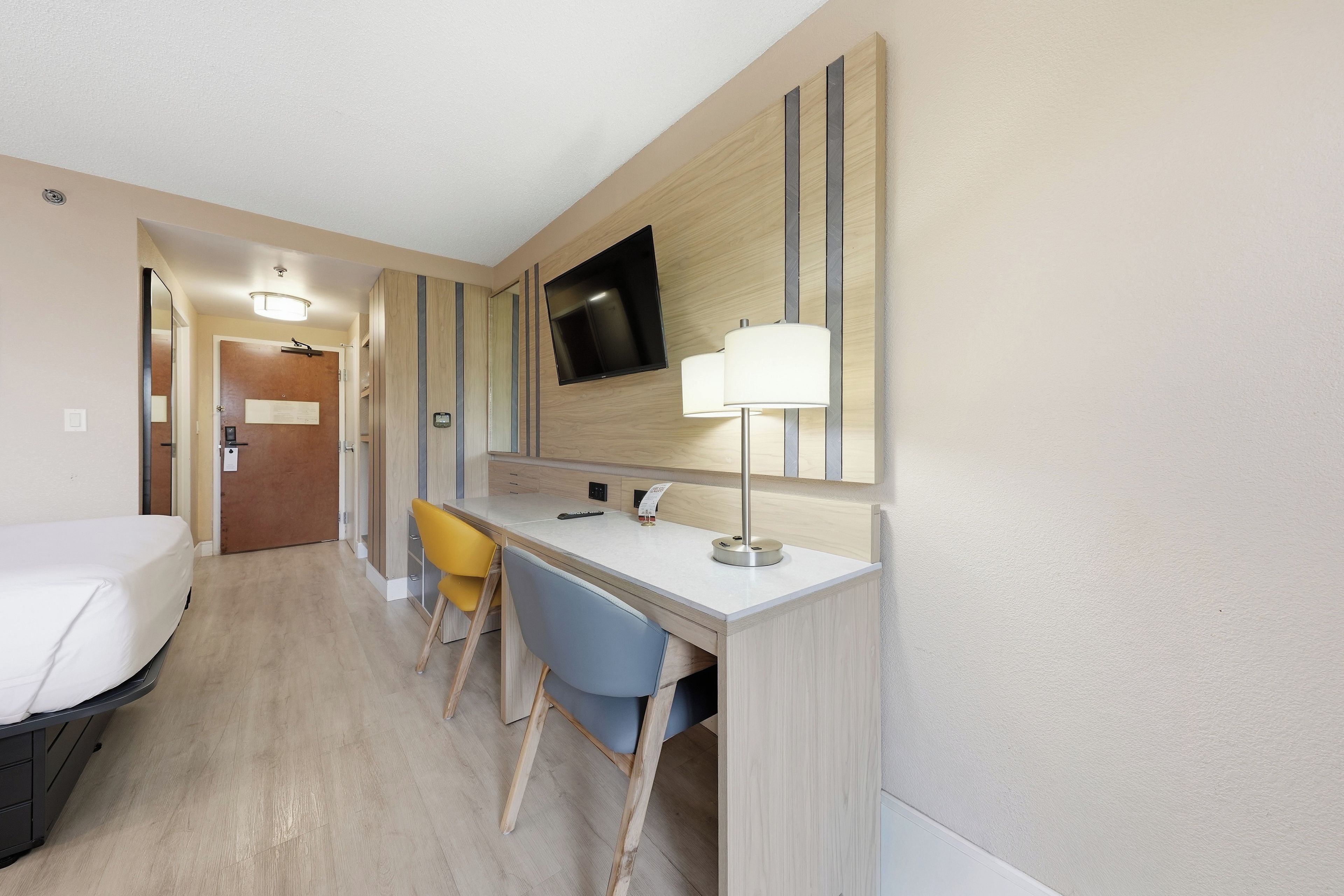 Ein Hotelzimmer mit einem Bett, einem Schreibtisch mit Stuhl, einem an der Wand befestigten Fernseher und einer Tür zu einem anderen Raum.
