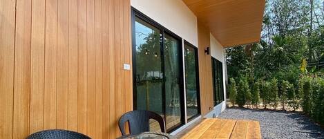 Una terraza de madera con dos sillas de mimbre y una mesa pequeña, techo de madera y una puerta de cristal que da a un jardín.