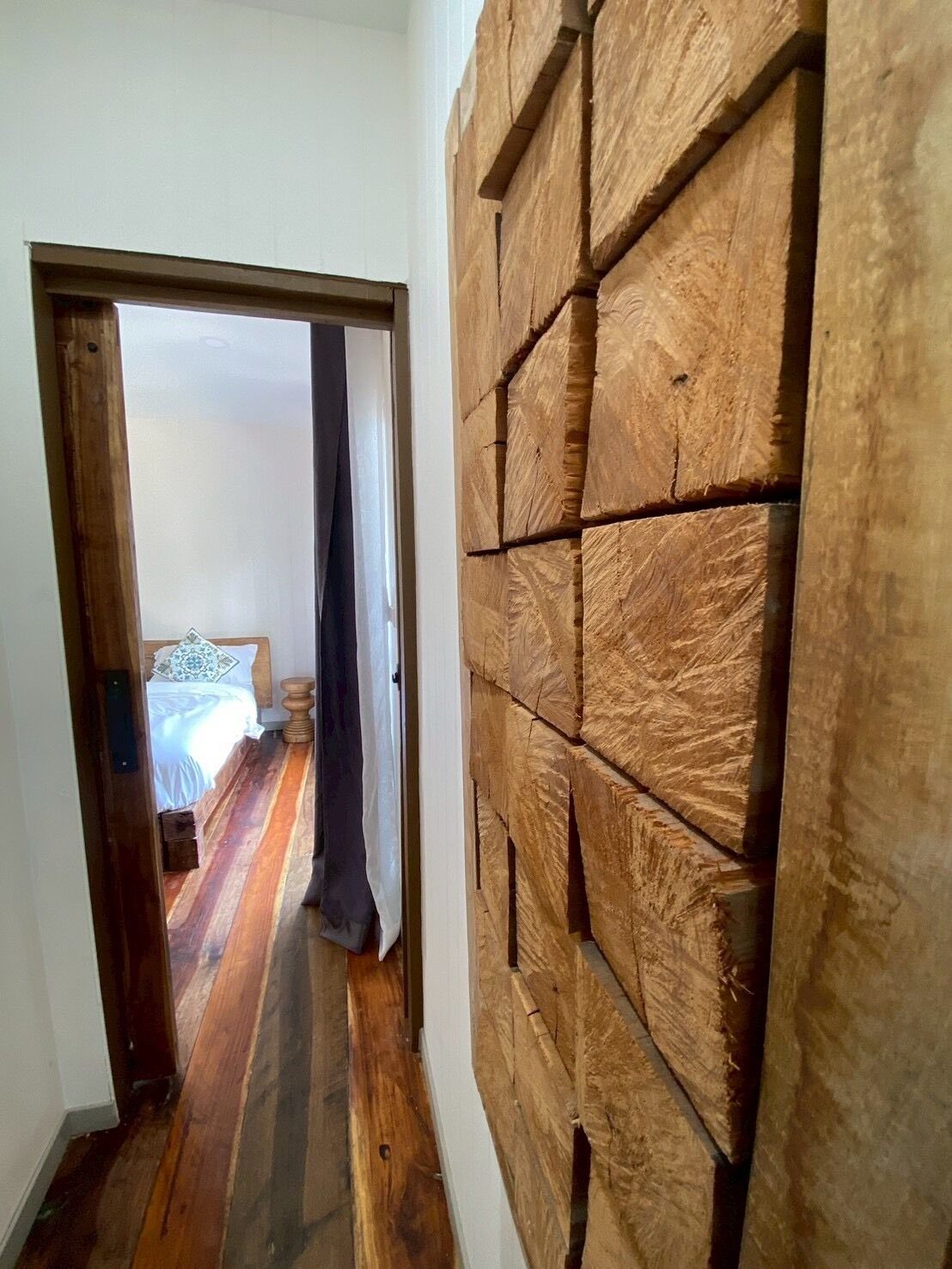 A hallway with wooden flooring leading to a bedroom with a bed and a nightstand.