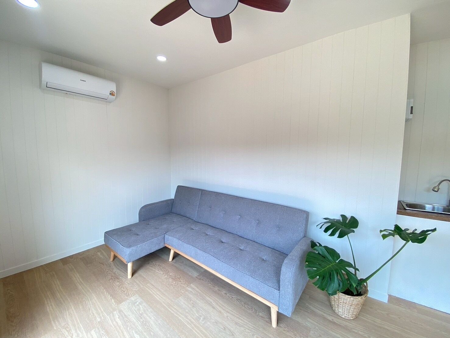 Une pièce avec un canapé d’angle gris, un ventilateur de plafond, un appareil de climatisation, un plancher en bois et une plante en pot.