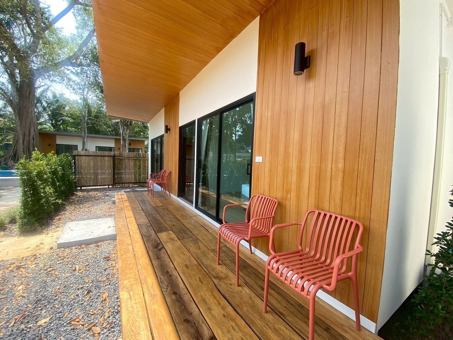 A covered outdoor seating area with wooden flooring, red chairs, and a wooden wall with a window.
