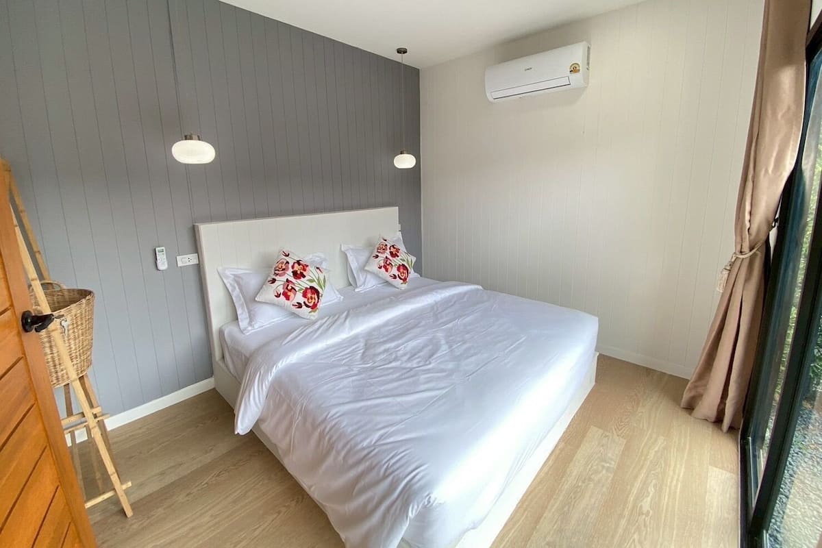A bedroom with a bed, a wooden side table, an air conditioning unit, and a window with curtains.